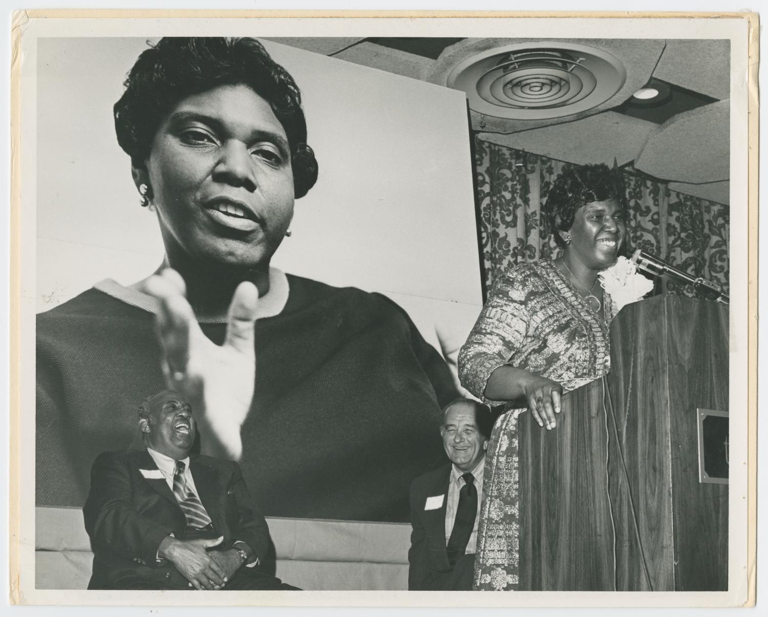 Barbara Jordan stands at a podium at a fundraiser on Oct. 21, 1971, at the Rice Hotel in Houston. An enlarged photograph of Jordan is visible in the background. Mack Hannah and Lyndon B. Johnson sit behind Jordan.