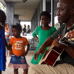Man playing a guitar with children surrounding 